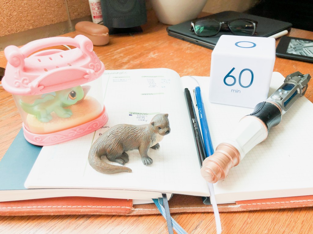 An open planner with two pens, a toy iguana, a toy otter, a timer and a replica of Dr. Who's sonic screwdriver